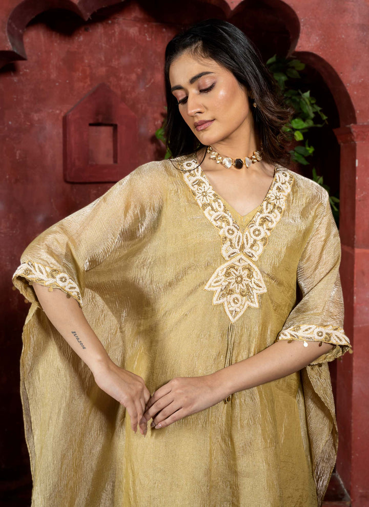 Woman wearing a gold traditional kaftan outfit with intricate white pearl embroidery against a red architectural background.