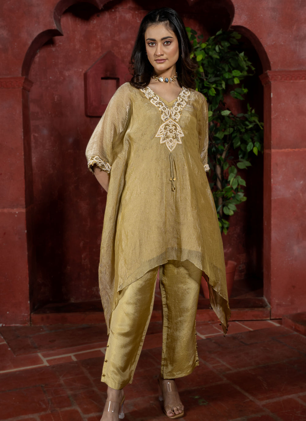 Woman wearing a gold traditional kaftan outfit with intricate white pearl embroidery against a red architectural background.