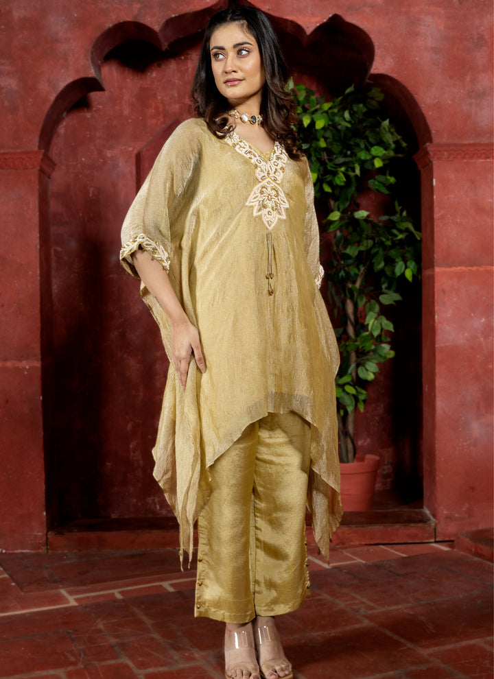 Woman wearing a gold traditional kaftan outfit with intricate white pearl embroidery against a red architectural background.