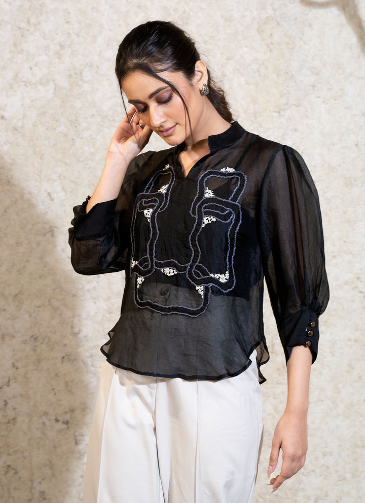 Woman wearing a black top with embroidery, against a textured white wall.