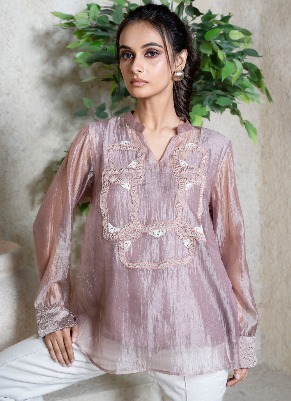 Woman wearing a light pink embroidered top with white pants, standing against a neutral background with greenery.