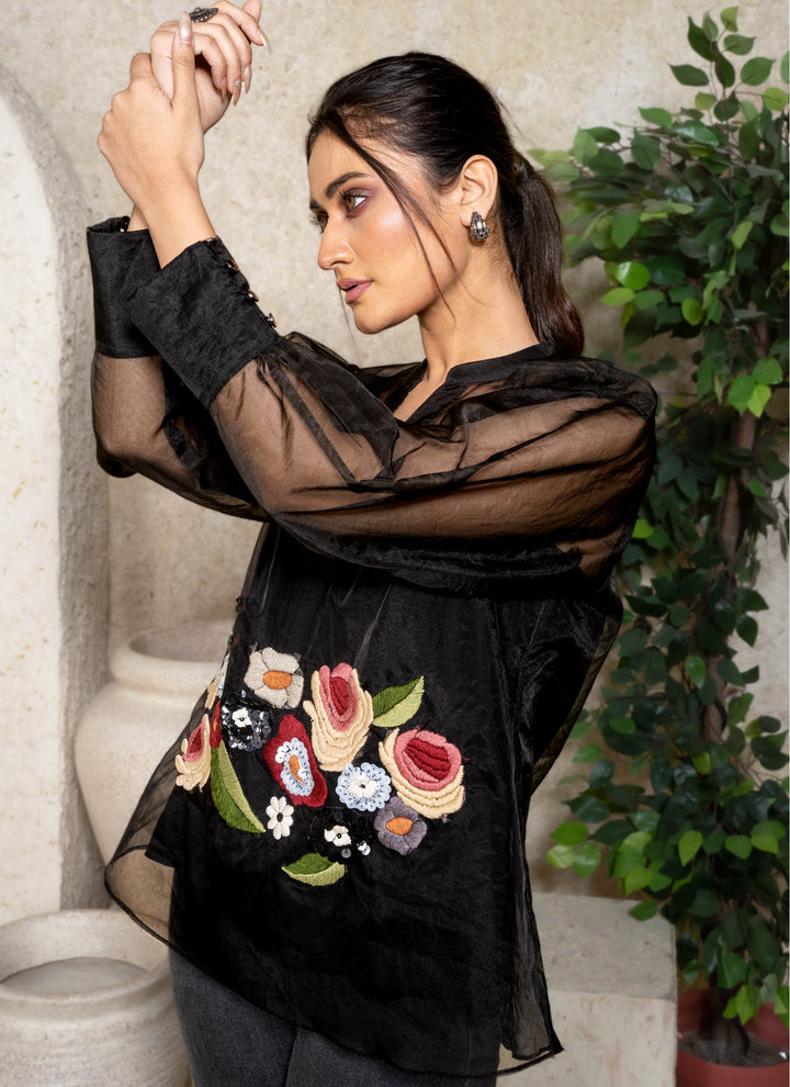 Woman wearing a black sheer top with floral embroidery in an indoor setting.