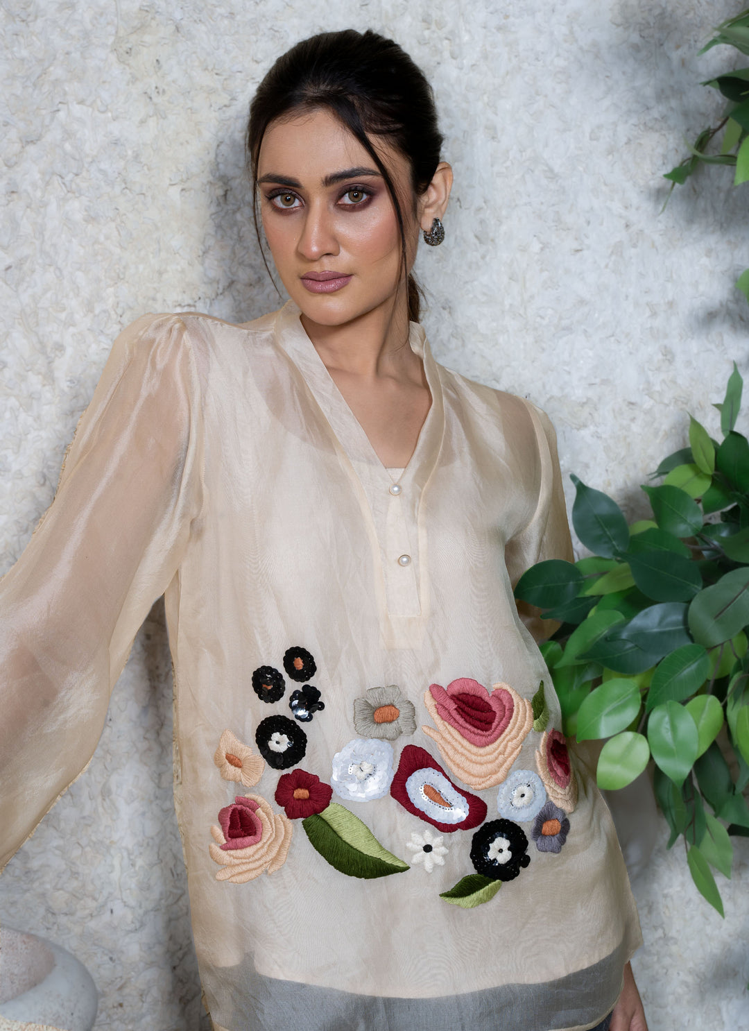 Woman wearing a cream top with floral embroidery against a textured white wall.