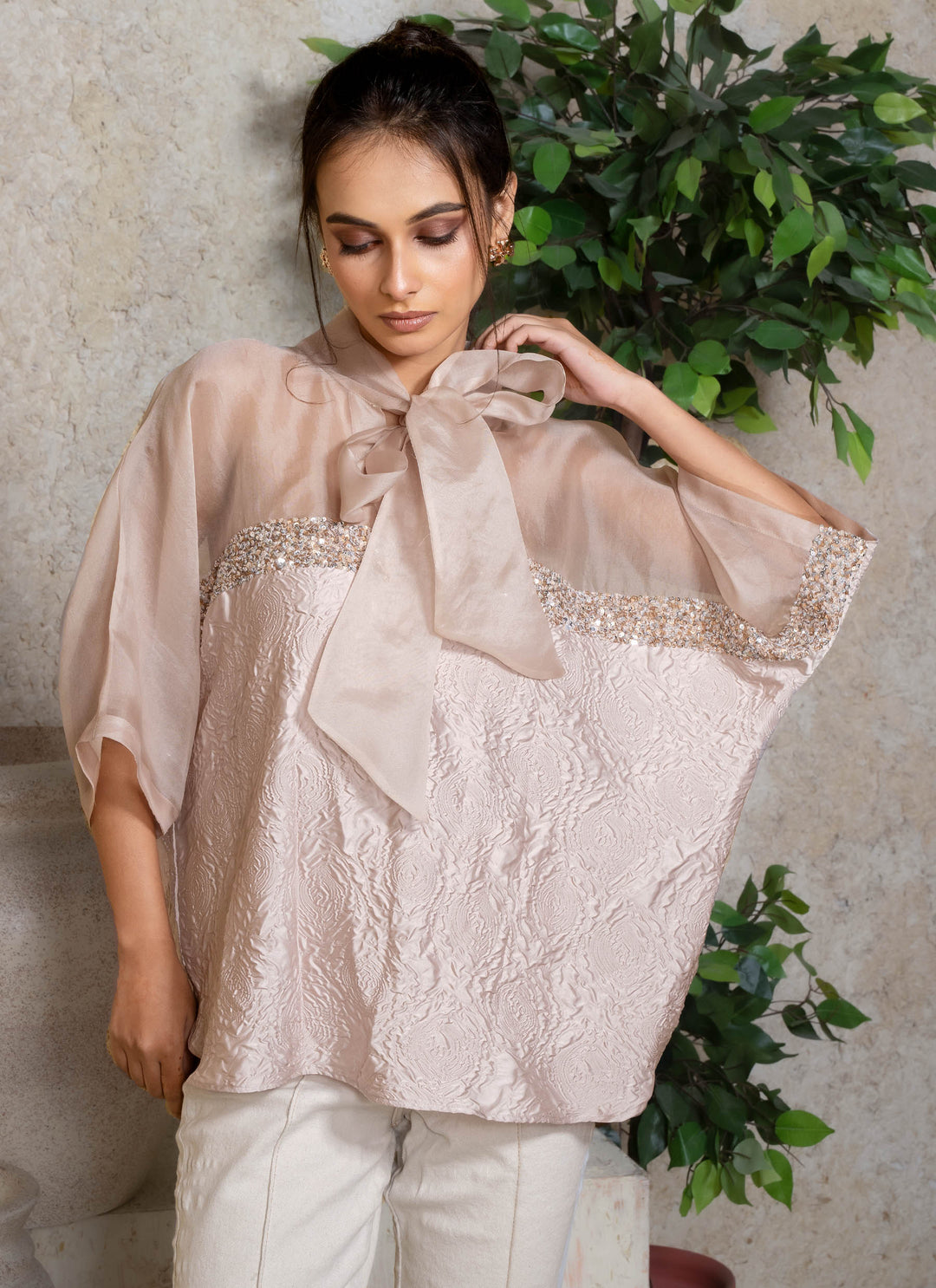 Woman wearing a light cream kaftan top with a large bow and sequins details, standing against a neutral background with greenery.