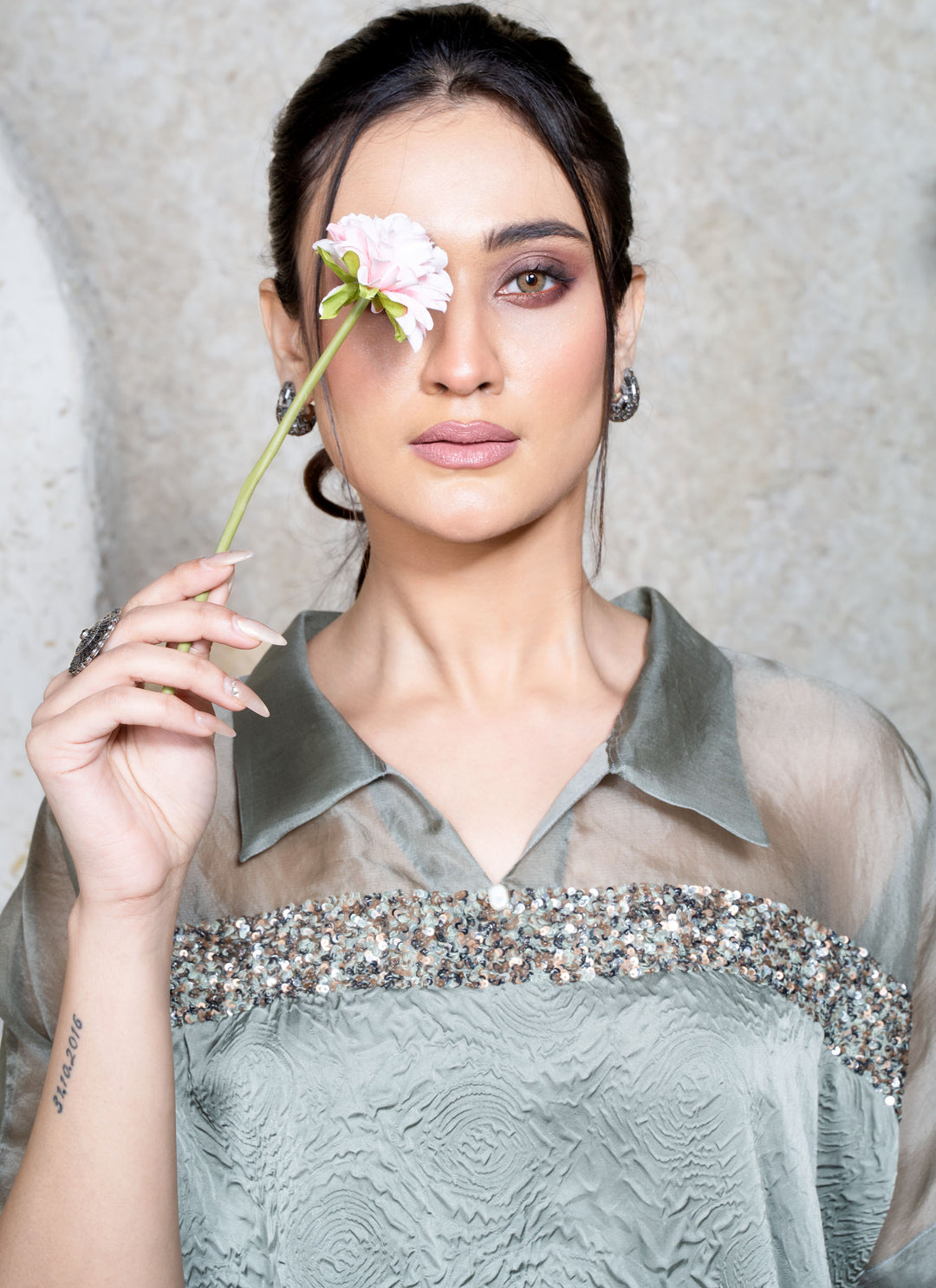 Woman holding a flower near her eye , wearing a  collar green top, with a light gray background