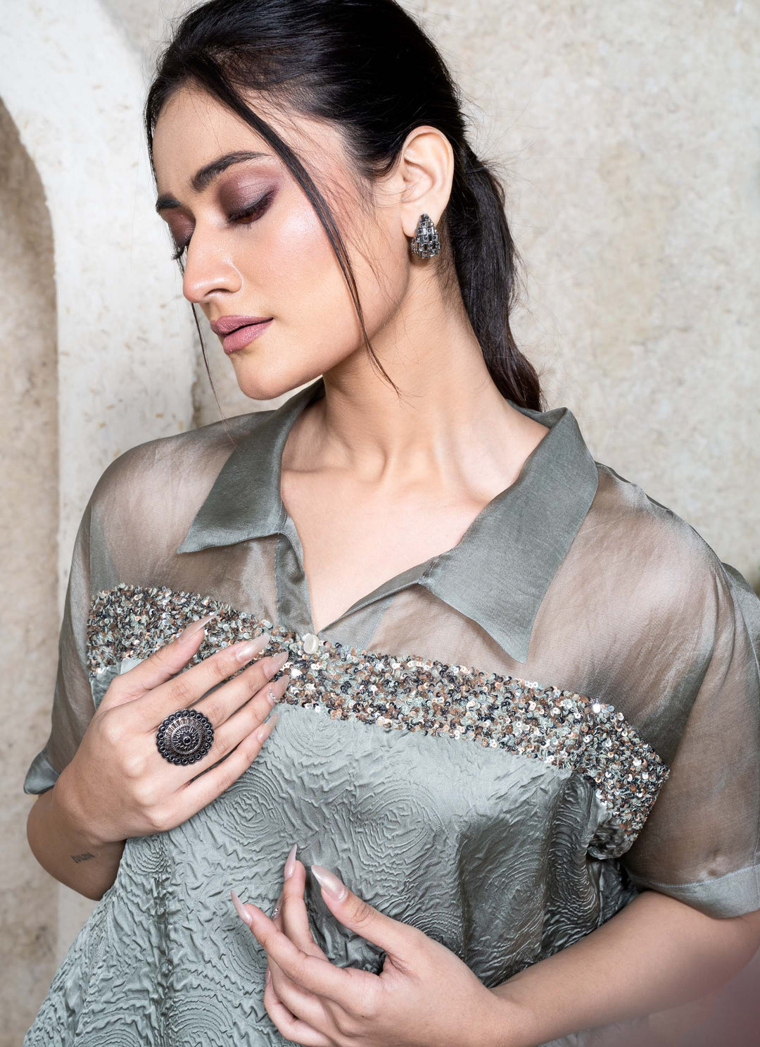 Woman wearing a green kaftan top with sequins work and white pants sitting against a neutral background.