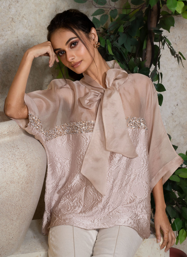 Woman wearing a light cream kaftan top with a large bow and sequins details, standing against a neutral background with greenery.