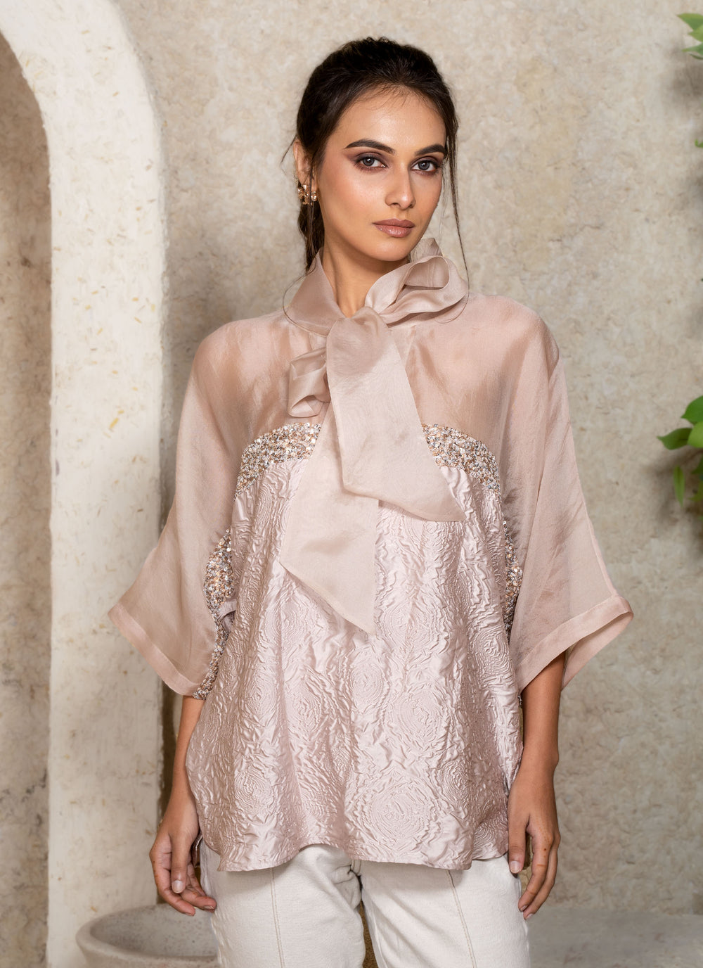 Woman wearing a light cream kaftan top with a large bow and sequins details, standing against a neutral background with greenery.