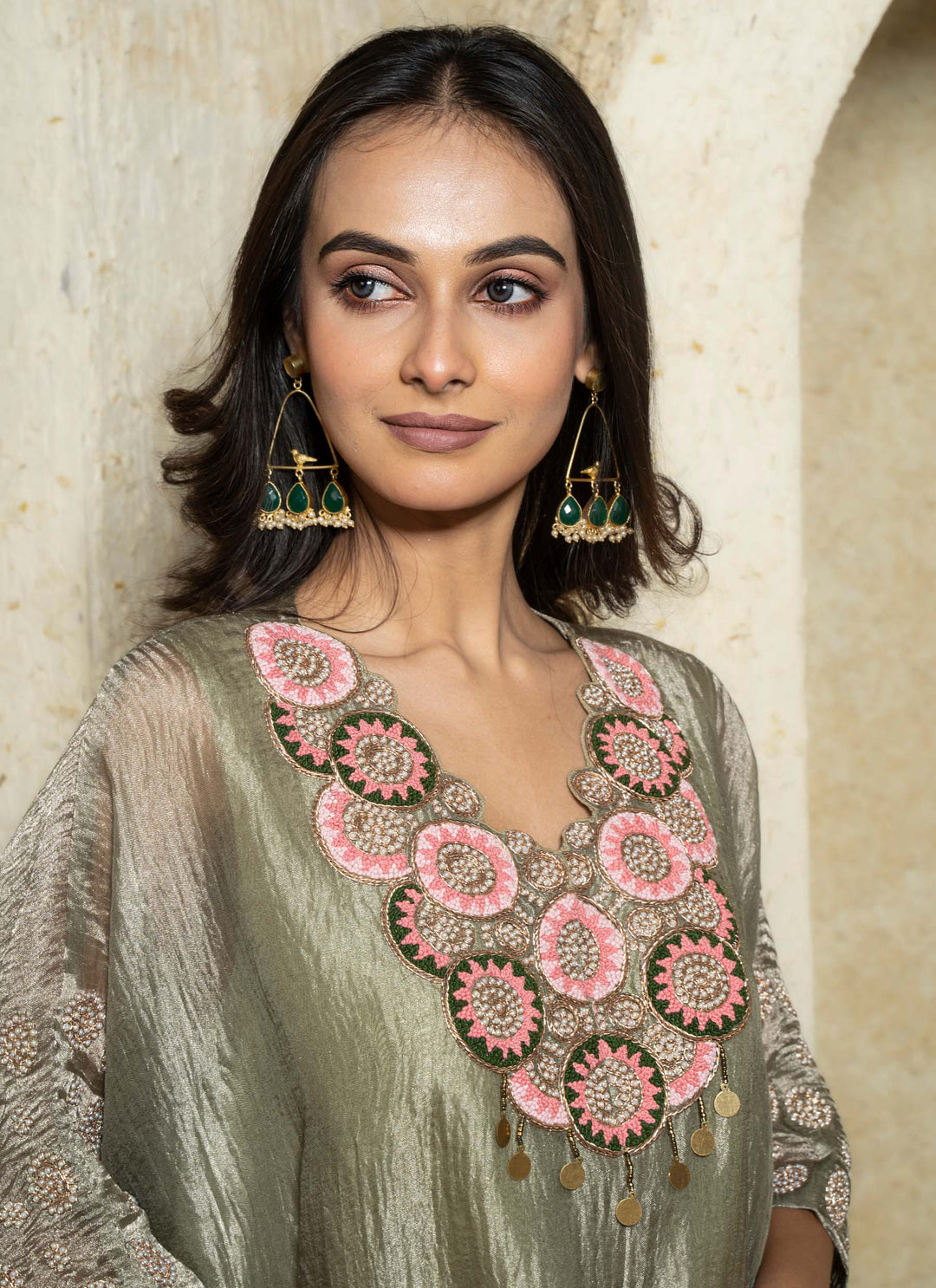 Woman wearing a traditional kaftan outfit with floral embroidery and green earrings against a beige wall.