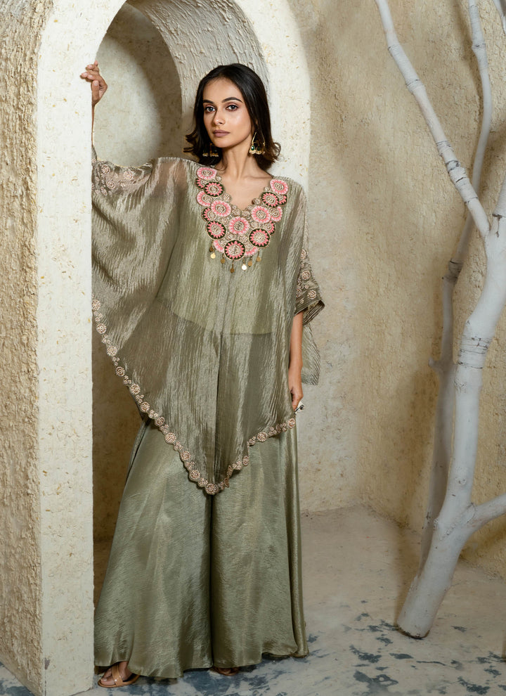 Woman wearing a long, light-colored kaftan outfit with floral patterns in a rustic setting.