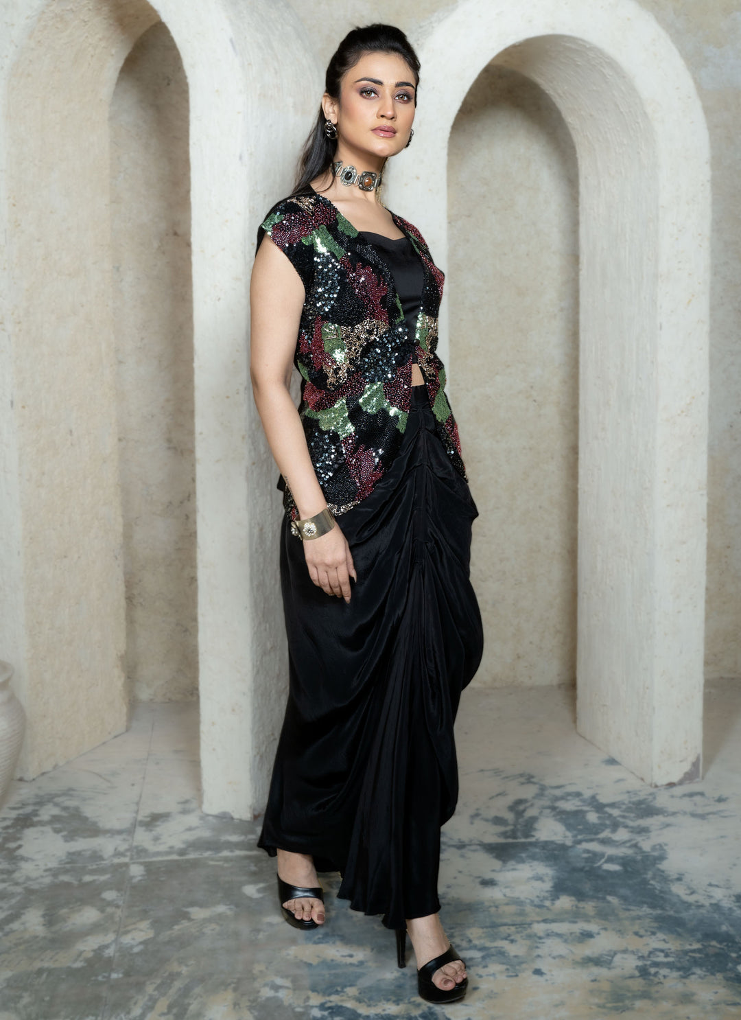 Woman in a black drape skirt with abstract sequins embroidered open jacket standing in an archway.