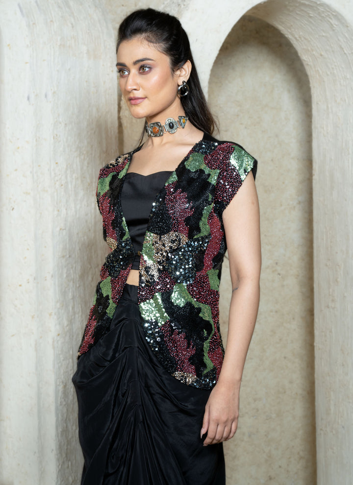 Woman in a black drape skirt with abstract sequins embroidered open jacket standing in an archway.