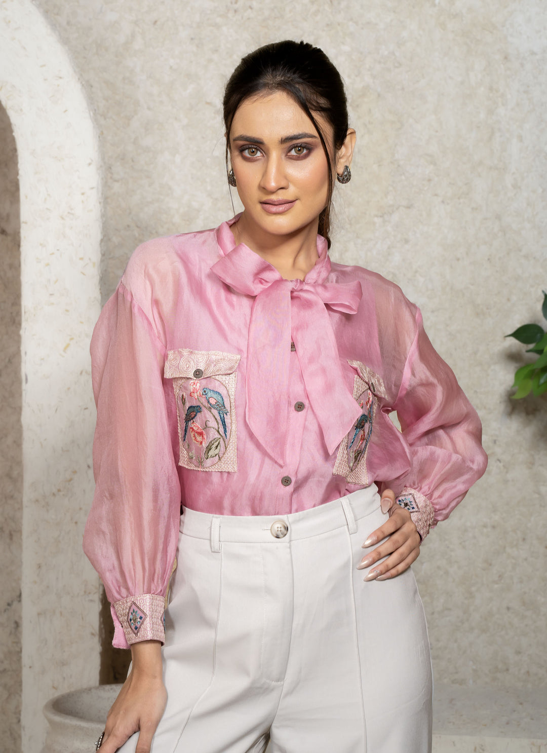 Woman wearing a pink shirt with embroidery and white pants in an indoor setting.