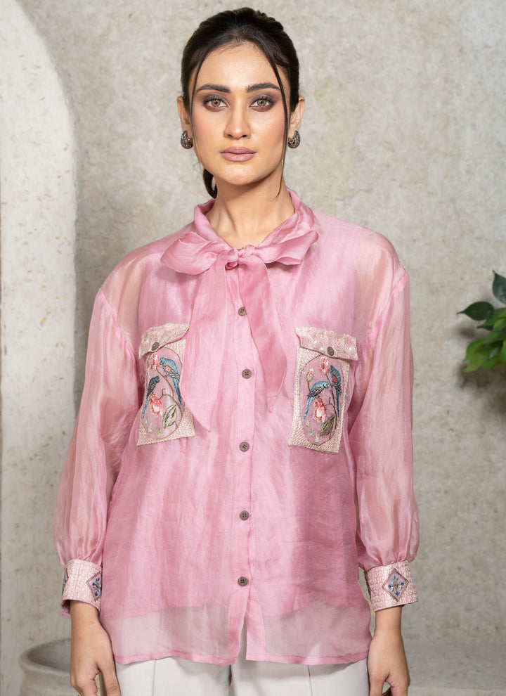 Woman wearing a pink shirt with embroidery, standing against a neutral background.