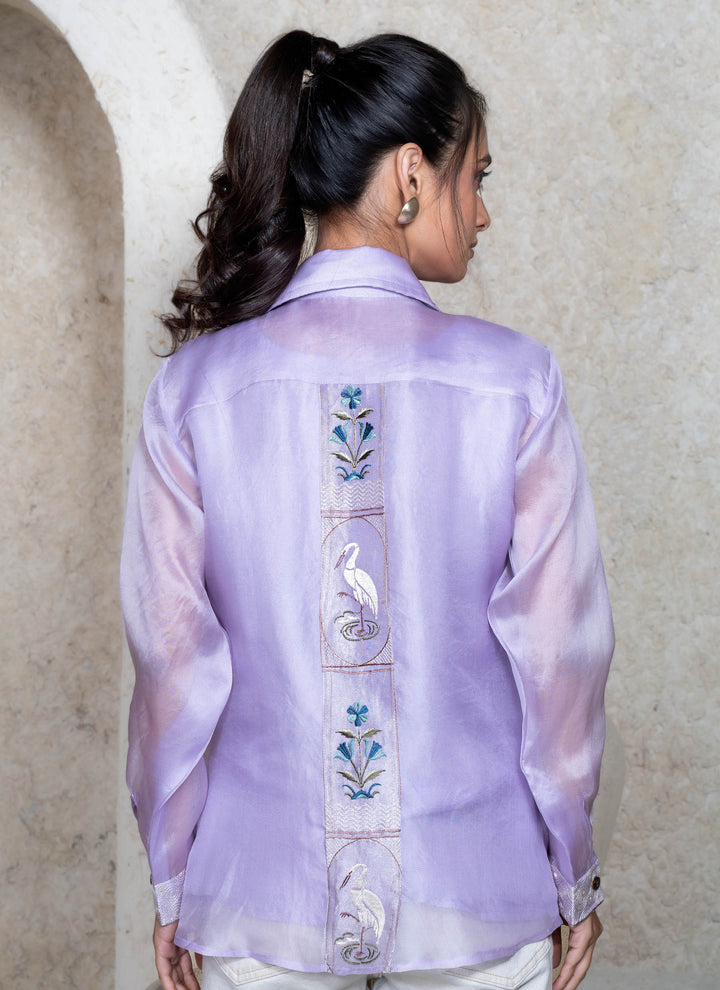 Woman wearing a lilac embroidered shirt with floral and bird designs, standing against a beige wall.