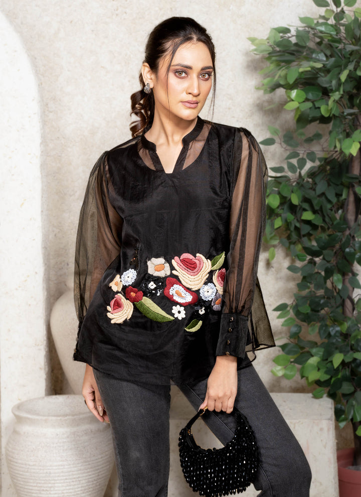 Woman wearing a black top with floral embroidery, walking against a neutral background.